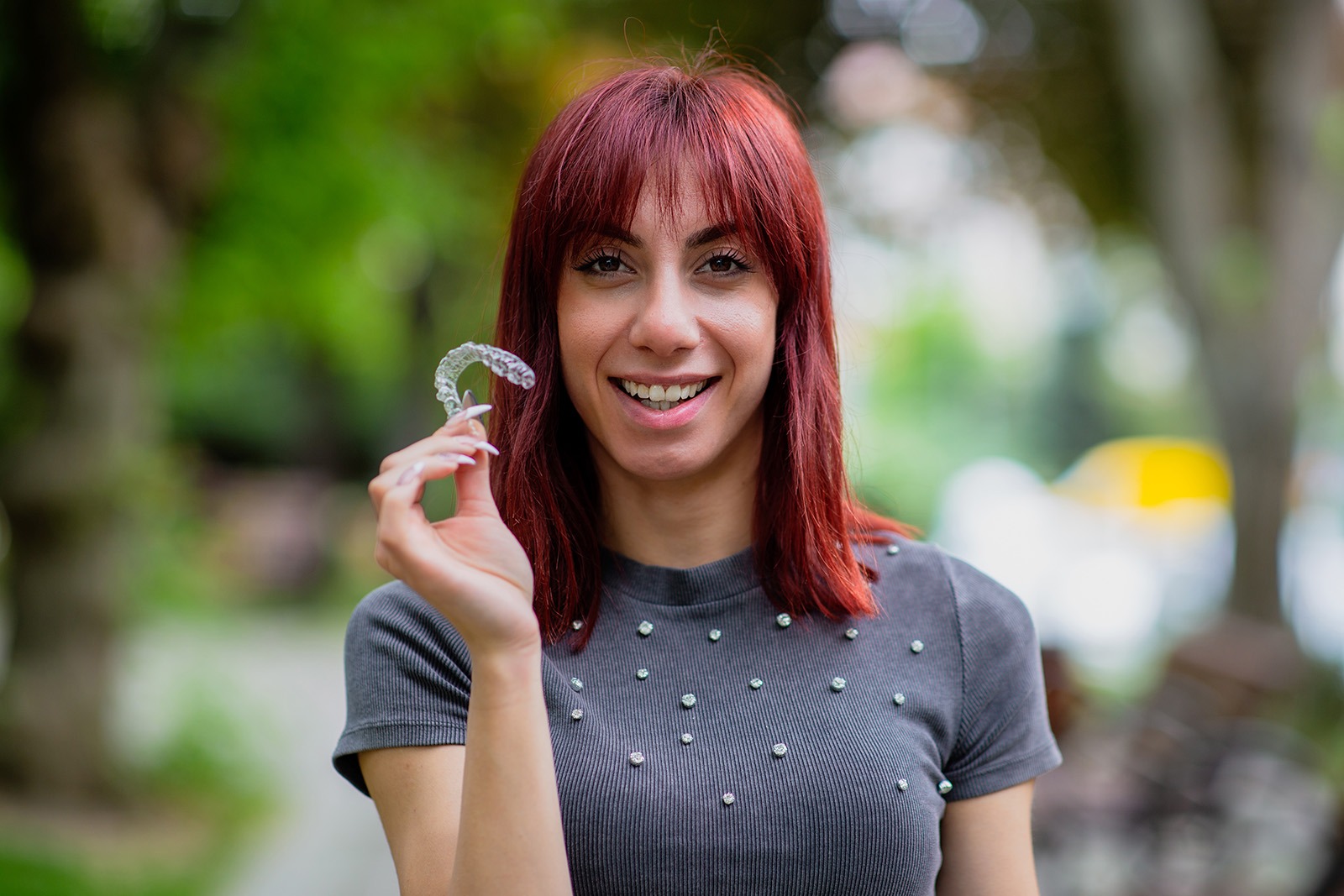 A smiling young woman with red hair is holding up a clear dental retainer